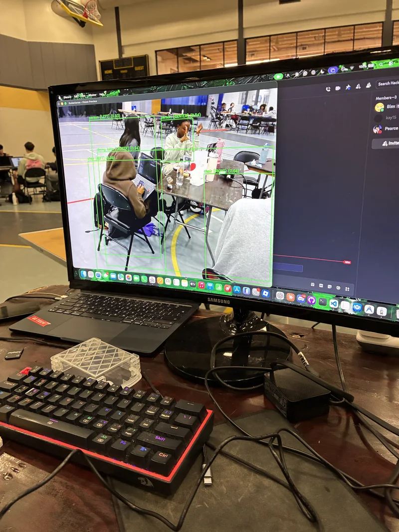 Photo of a monitor running the early GuideSense vision preview, with green detection boxes around people and chairs in the hackathon room.